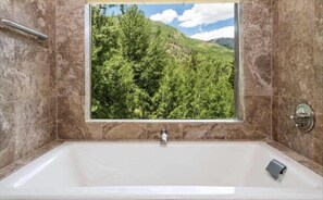 Un bagno con una grande vasca, una finestra vista foresta e pareti in marmo.