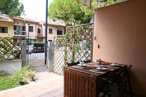 A wooden table set for dining with chairs, a metal gate, and a view of neighboring buildings. - S255 - Sirolo, new three-room apartment a stone's throw from the center (Sirolo)