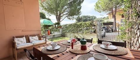 An outdoor dining area with a wooden table set for two, surrounded by chairs and a bench.