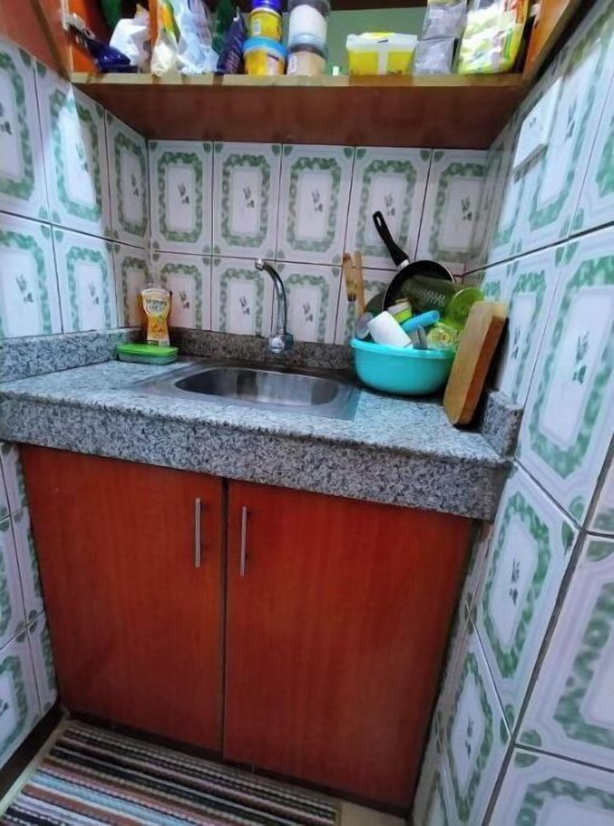 Une petite cuisine avec un comptoir en granit, un évier et un meuble en bois. On y trouve divers ustensiles de cuisine sur le comptoir, notamment un saladier vert, une poêle et une planche à découper.