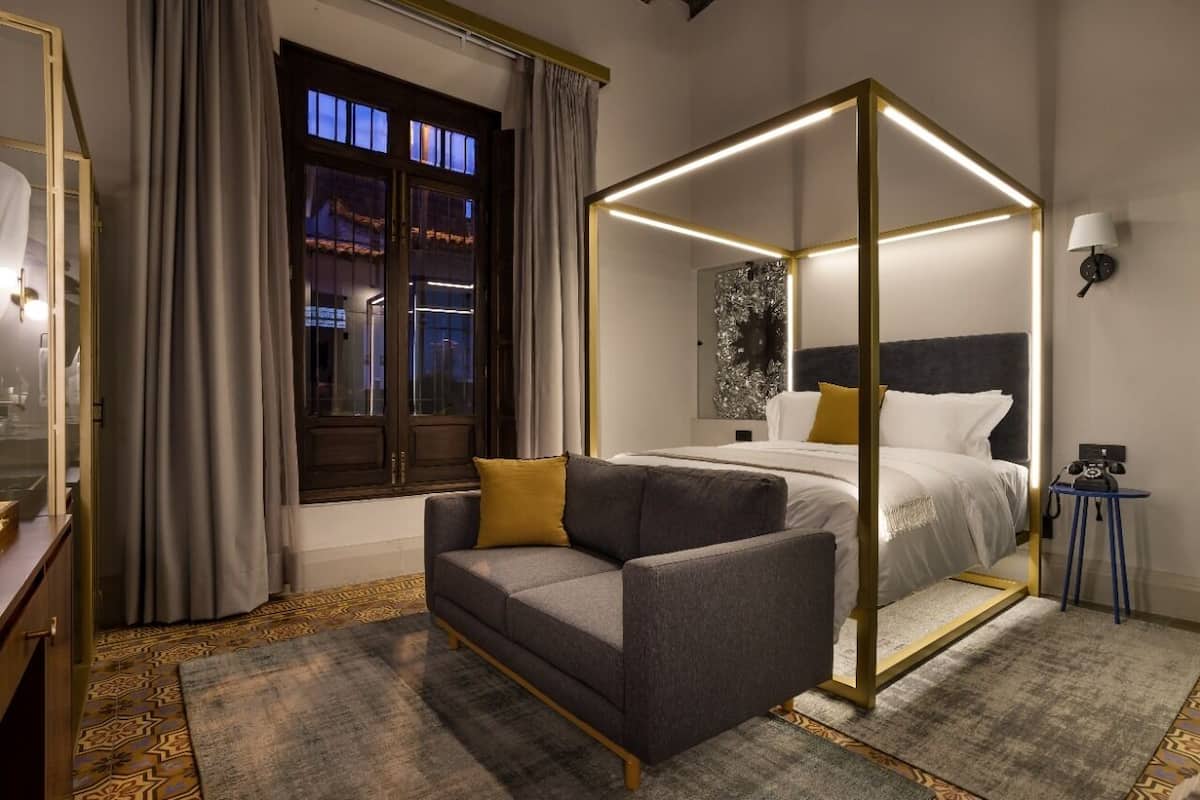 A modern hotel room with a four-poster bed, a sofa, a small table, and a window with curtains.