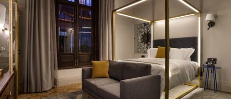 Una habitación de hotel moderna con una cama con dosel, un sofá, una mesita y un ventanal con cortinas.