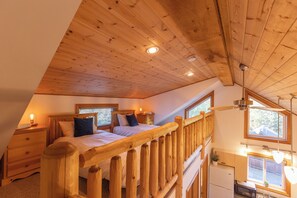 Une chambre de style « loft », avec un plafond en bois, deux lits, une table de chevet, une commode, une fenêtre et un coin cuisine équipé d’un réfrigérateur et d’un four à micro-ondes.