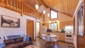 Ein gemütliches Wohnzimmer mit Sofa, Couchtisch und einem Gemälde eines Sees. Angrenzend an die Küche befindet sich ein Essbereich mit Tisch und Stühlen, inklusive Kühlschrank, Mikrowelle und Herd.