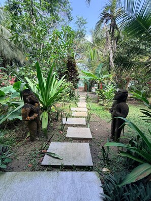 A stone pathway leading through a lush garden with tropical plants and two stone statues on either side.