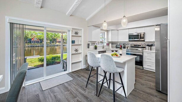 A modern kitchen with white cabinetry, a central island with bar stools, a large window with a view of a lake, and a dining area with a bench. - NEW Heated Salt Pool & Spa on Waterfront Canal (Tampa)