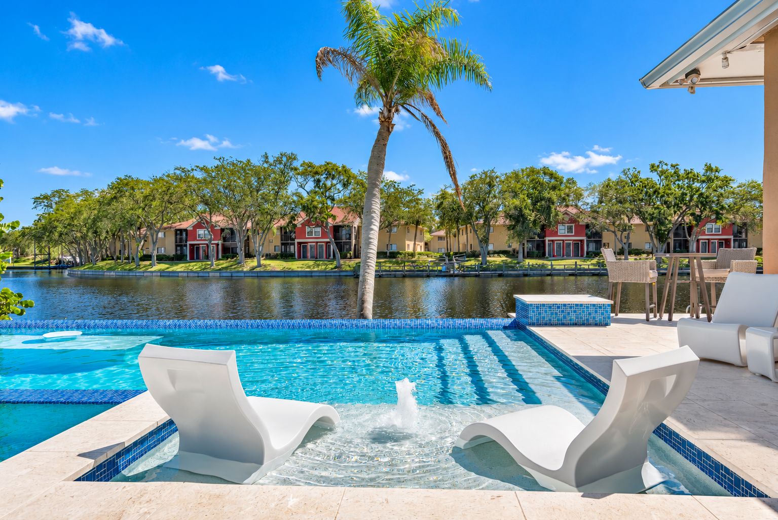 Una piscina con fuente, sillas de descanso y vistas a una zona residencial con casas y árboles.