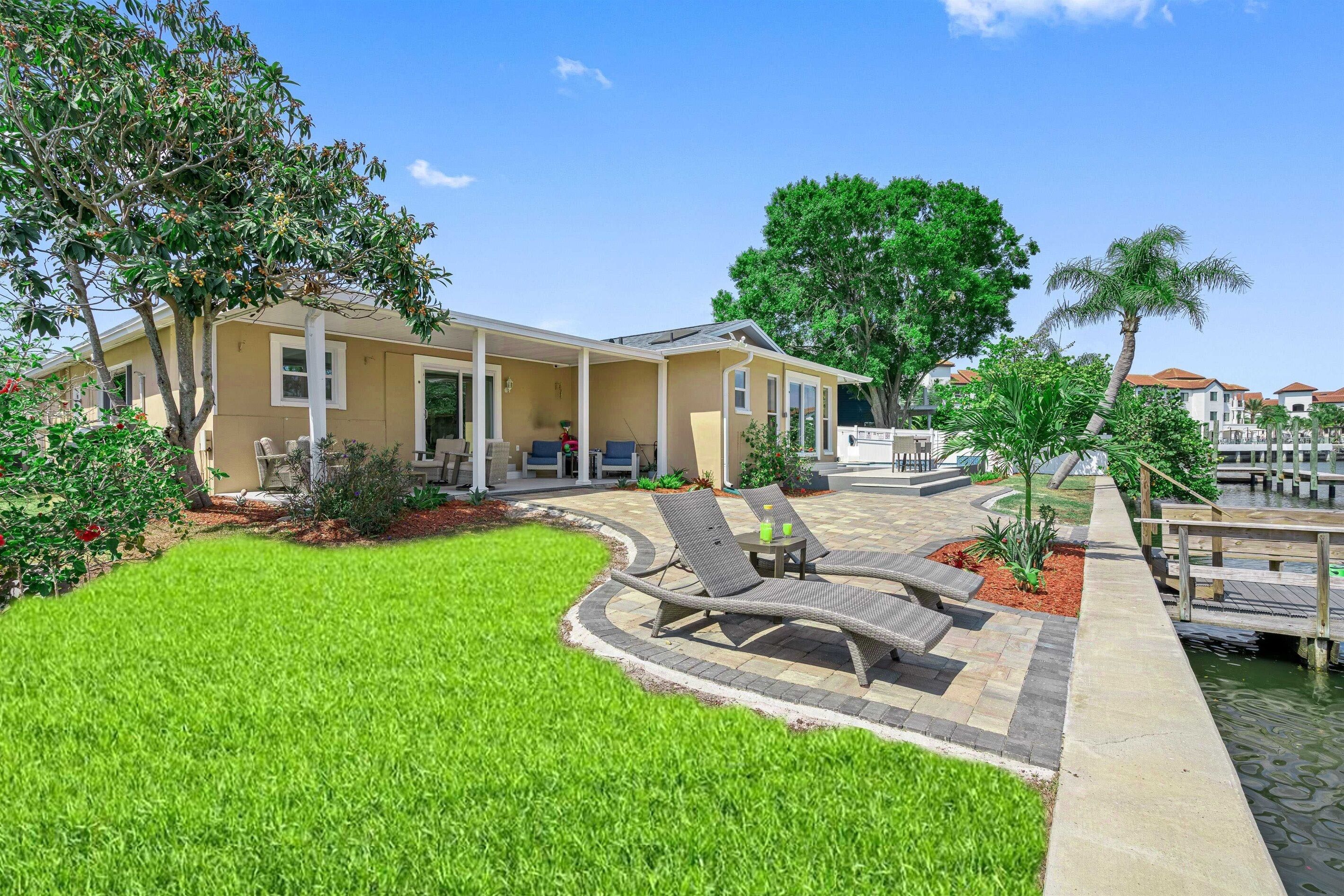 A residential property with a patio area featuring lounge chairs and a small table, surrounded by a well-maintained lawn and a water feature.