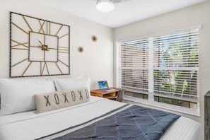 A bedroom with a bed, a wall clock, a desk with a laptop, and a window with blinds.