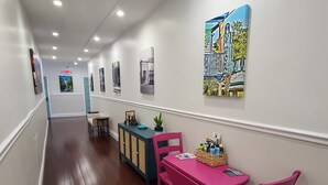 A hallway with a wooden floor, a pink table with chairs, a blue cabinet, and wall art.