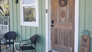 Une porte en bois avec une pancarte indiquant «Bienvenue», une véranda avec deux chaises et une fenêtre avec des stores.