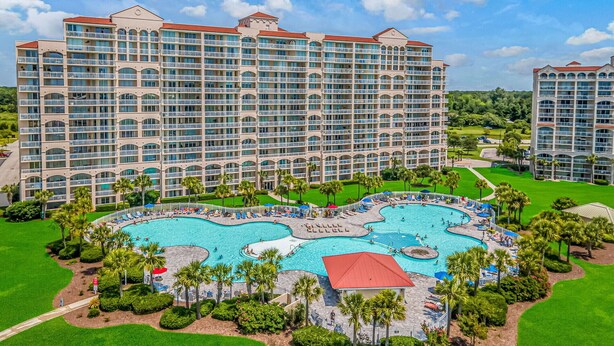 A large hotel with multiple balconies and a central swimming pool area surrounded by palm trees and sun loungers.