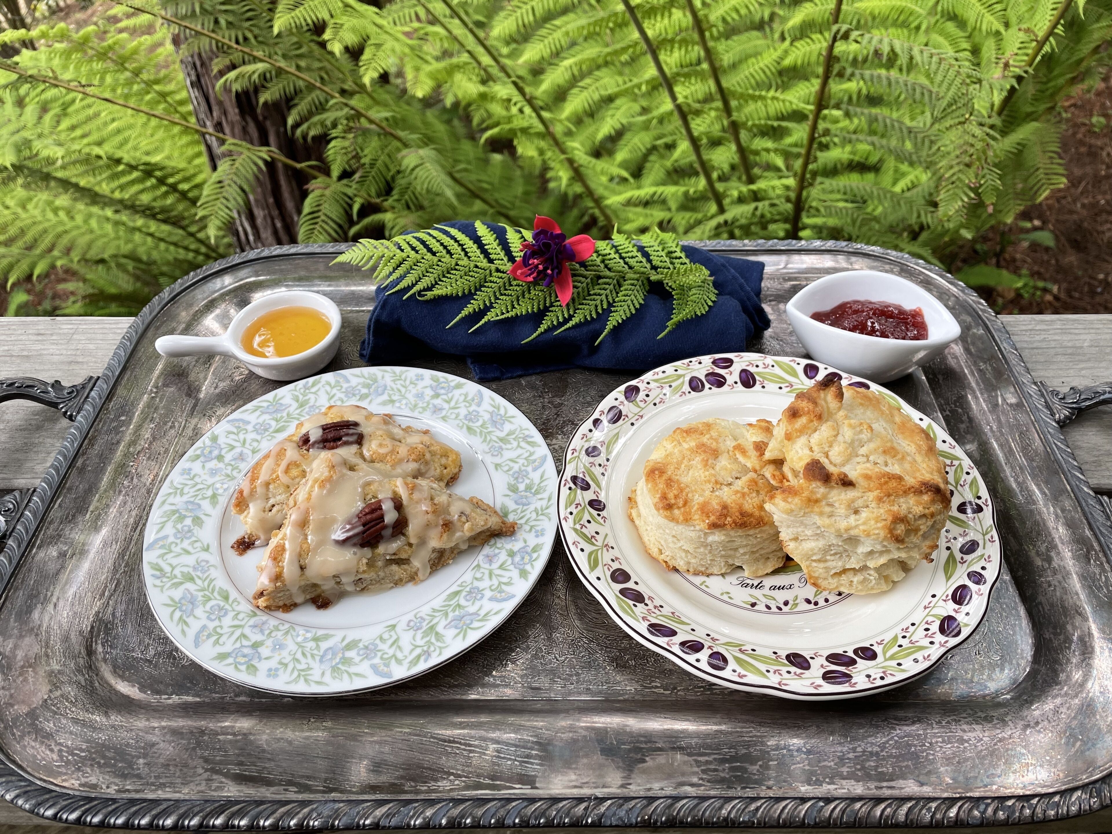 Uma bandeja com duas travessas de comida, uma com scones e outra com biscoitos, acompanhadas por uma pequena tigela de geleia e uma de creme.