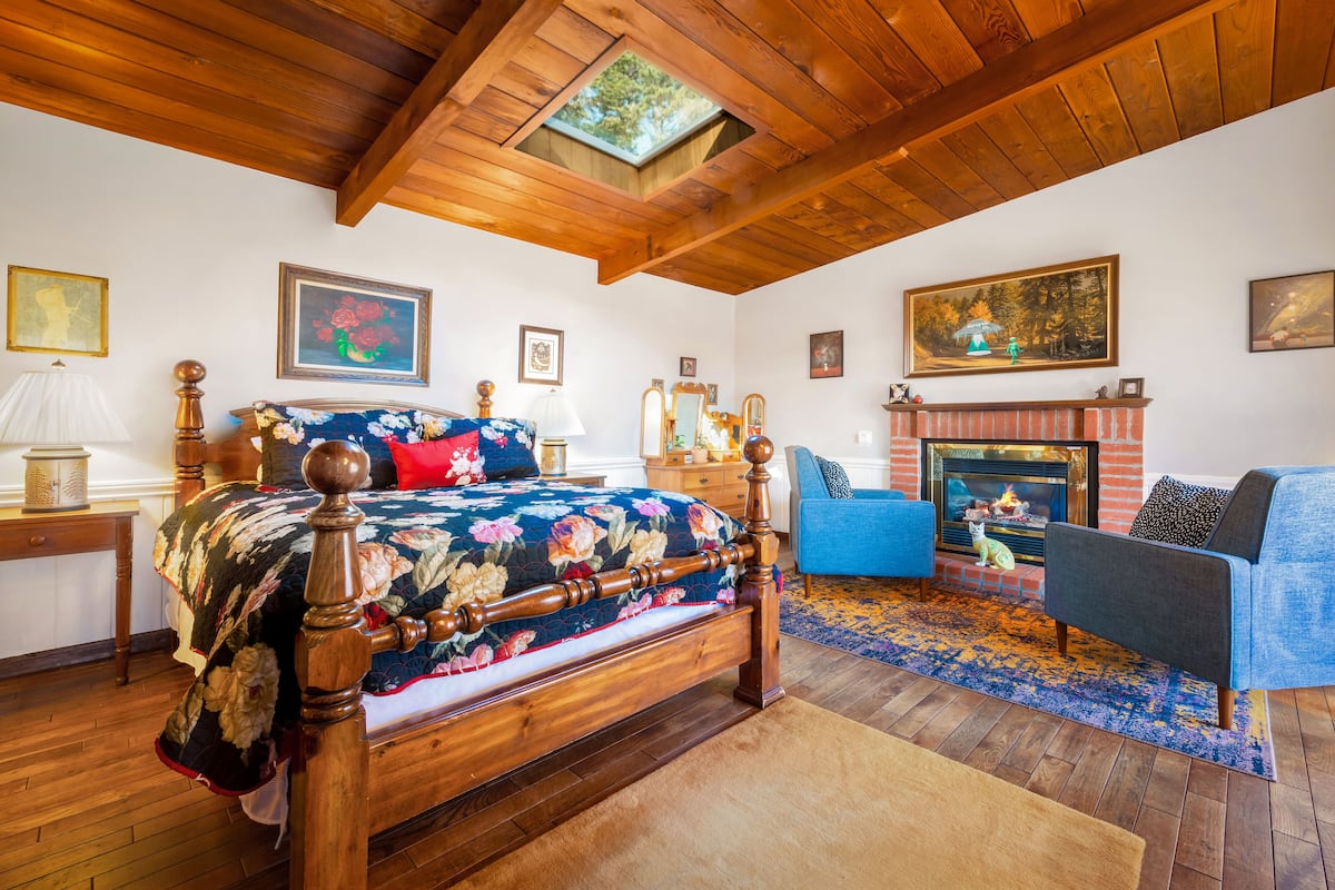Un dormitorio con techo de madera, una cama con colcha estampada, un sillón azul, chimenea y cuadros en las paredes.