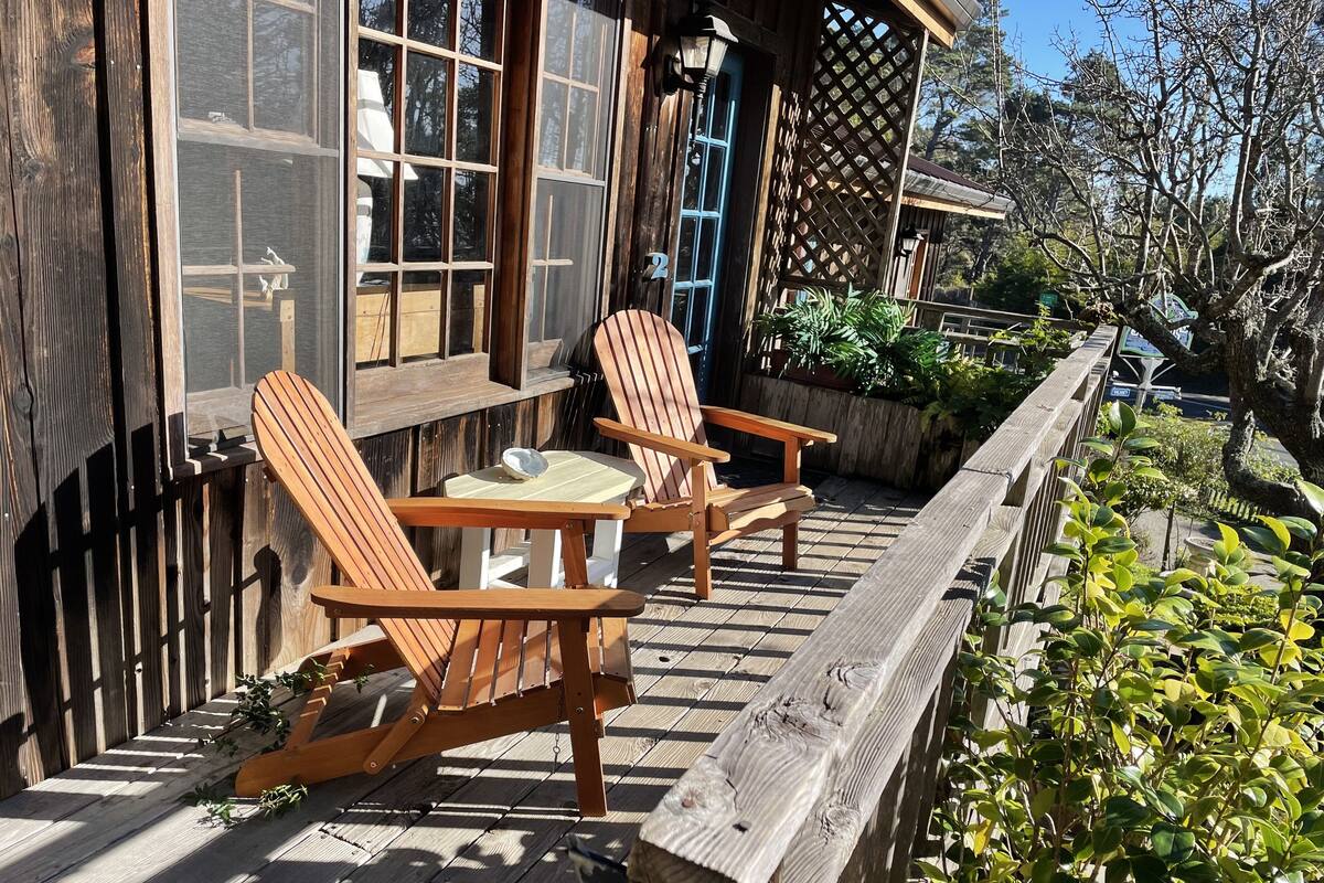 Una terraza de madera con dos sillas Adirondack y una pequeña mesa, con vista a un jardín que incluye una cerca de madera y una estructura de celosía.