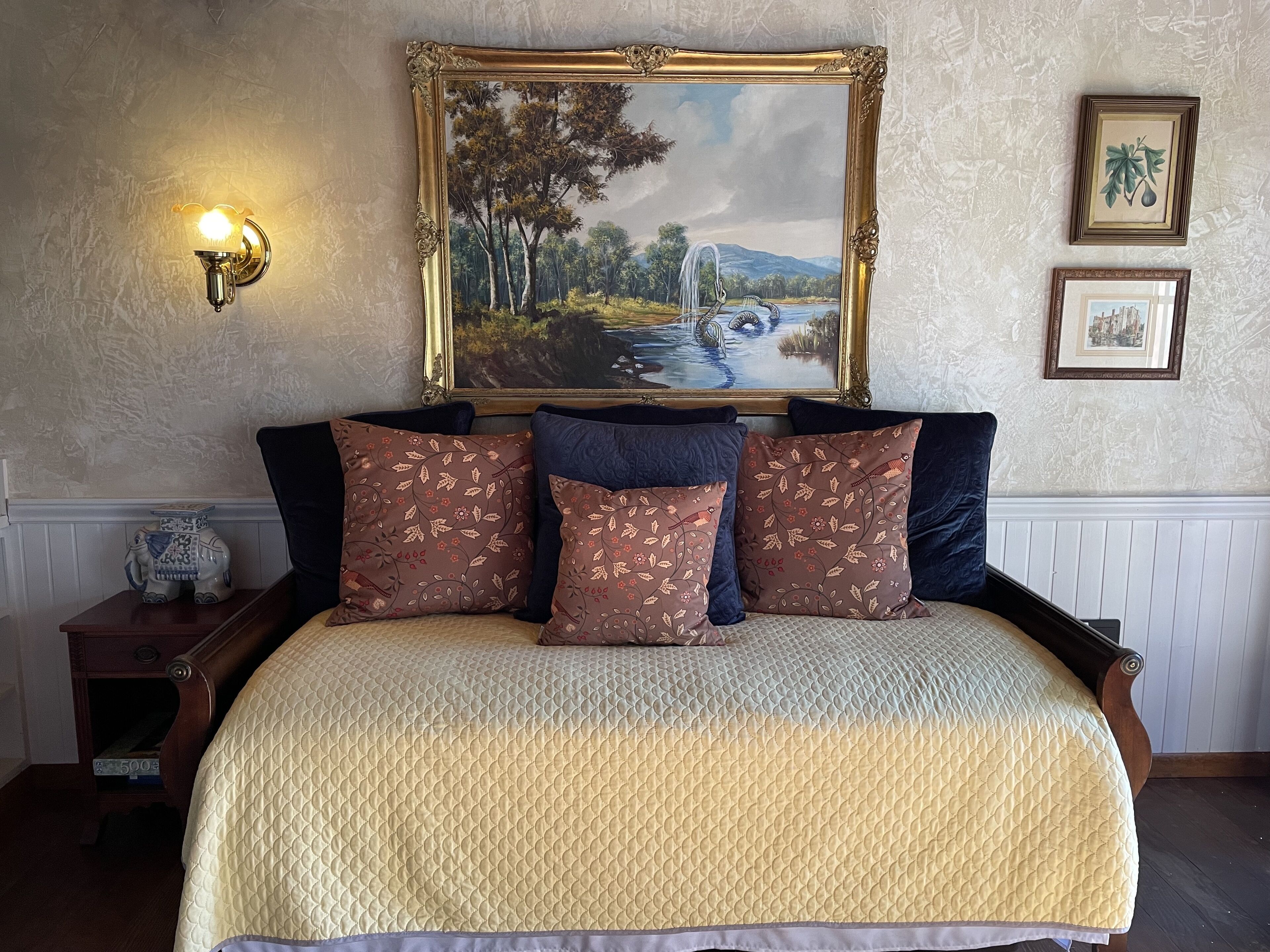 Une chambre à coucher avec un grand lit, un tableau représentant un paysage, une lampe fixée au mur et une petite table sur laquelle est posé un objet décoratif.