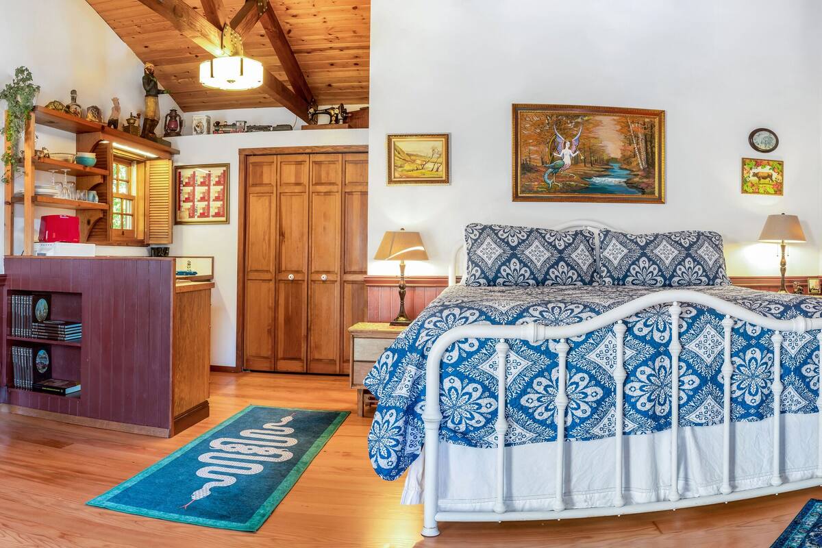 Un cuarto acogedor con una mesa de madera, sillas, una cama con cubrecama estampado y un cuadro grande sobre la cama.