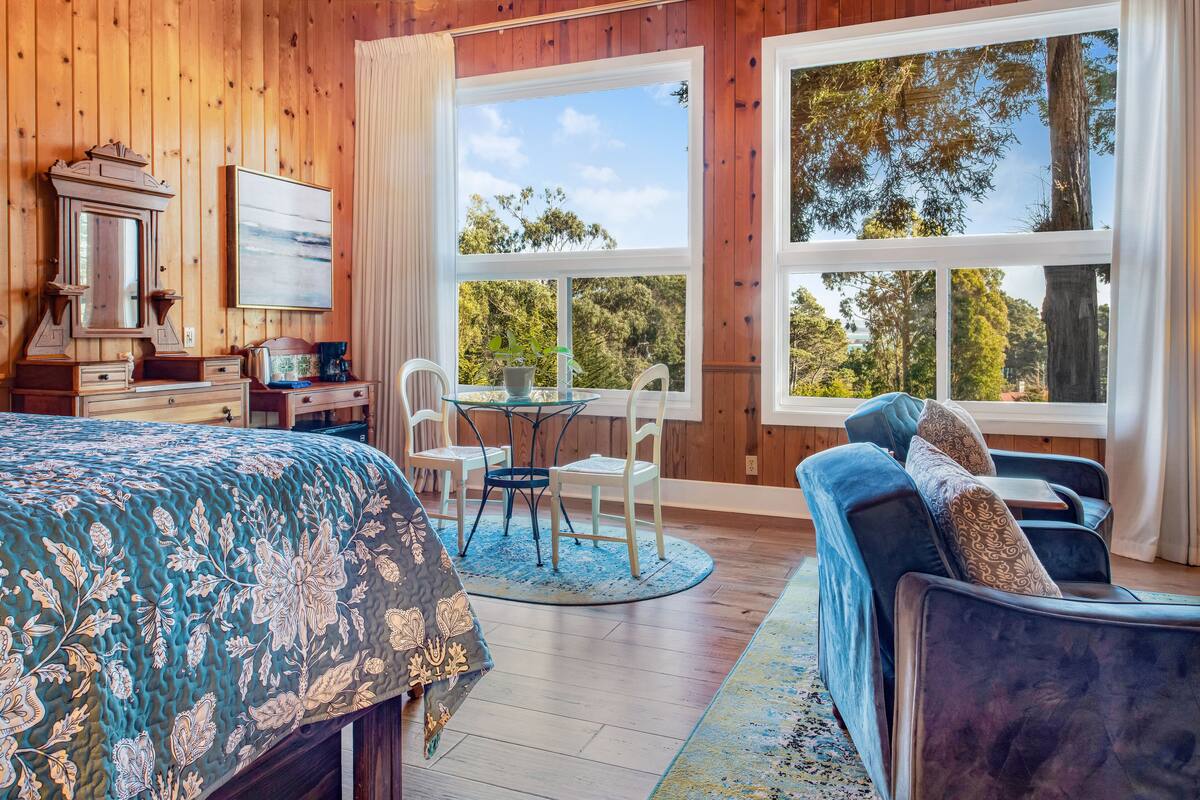 Una habitación con una cama, un escritorio con espejo, una silla, un sofá y dos ventanas con vistas a árboles.