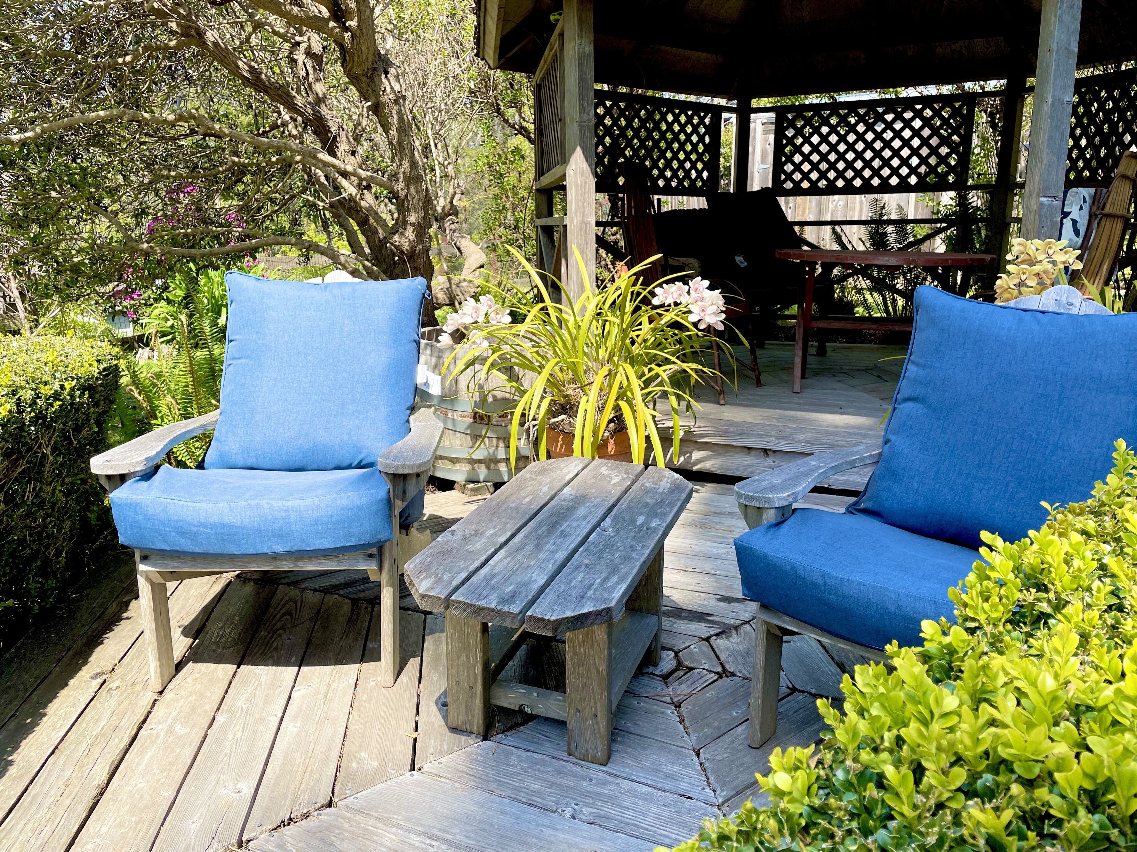 A wooden deck with two blue cushioned chairs and a small table, surrounded by greenery and a wooden pergola.