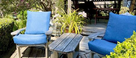 A wooden deck with two blue cushioned chairs and a small table, surrounded by greenery and a wooden pergola.