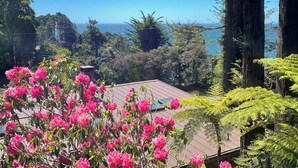 A view of a house with a wooden deck and pink flowers in the foreground, surrounded by tall trees and a clear blue sky. - Rendezvous Mendocino (Gualala)