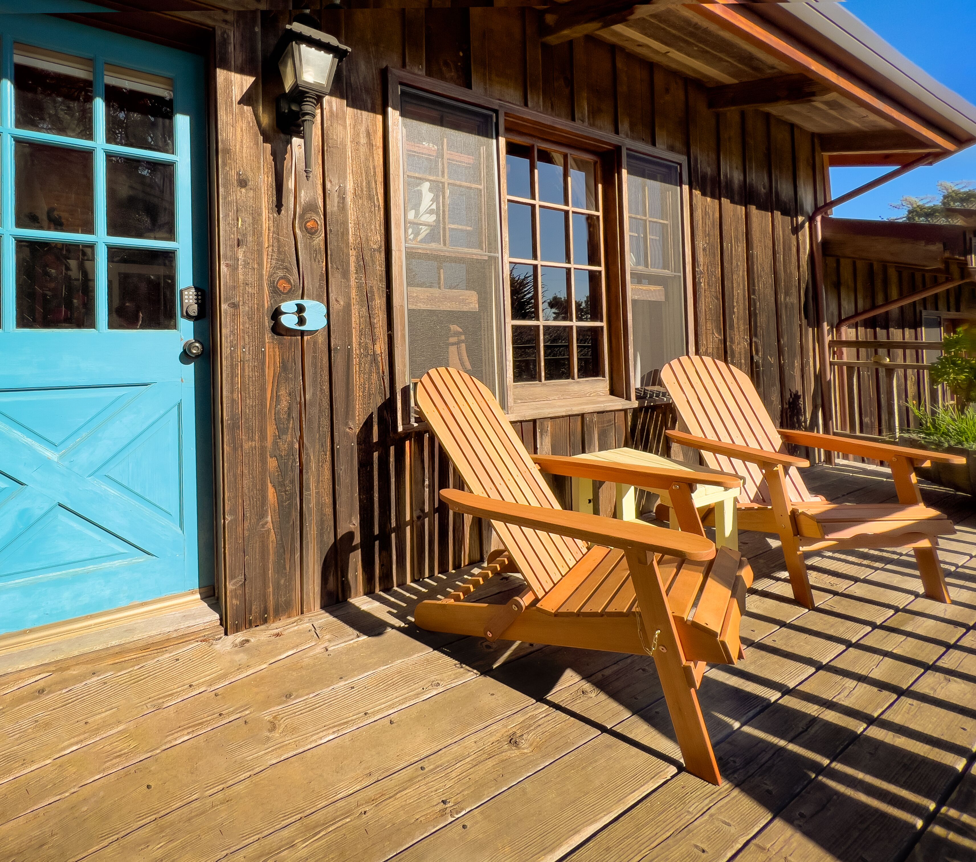 A wooden cabin with a blue door, two Adirondack chairs, and a wooden deck.
