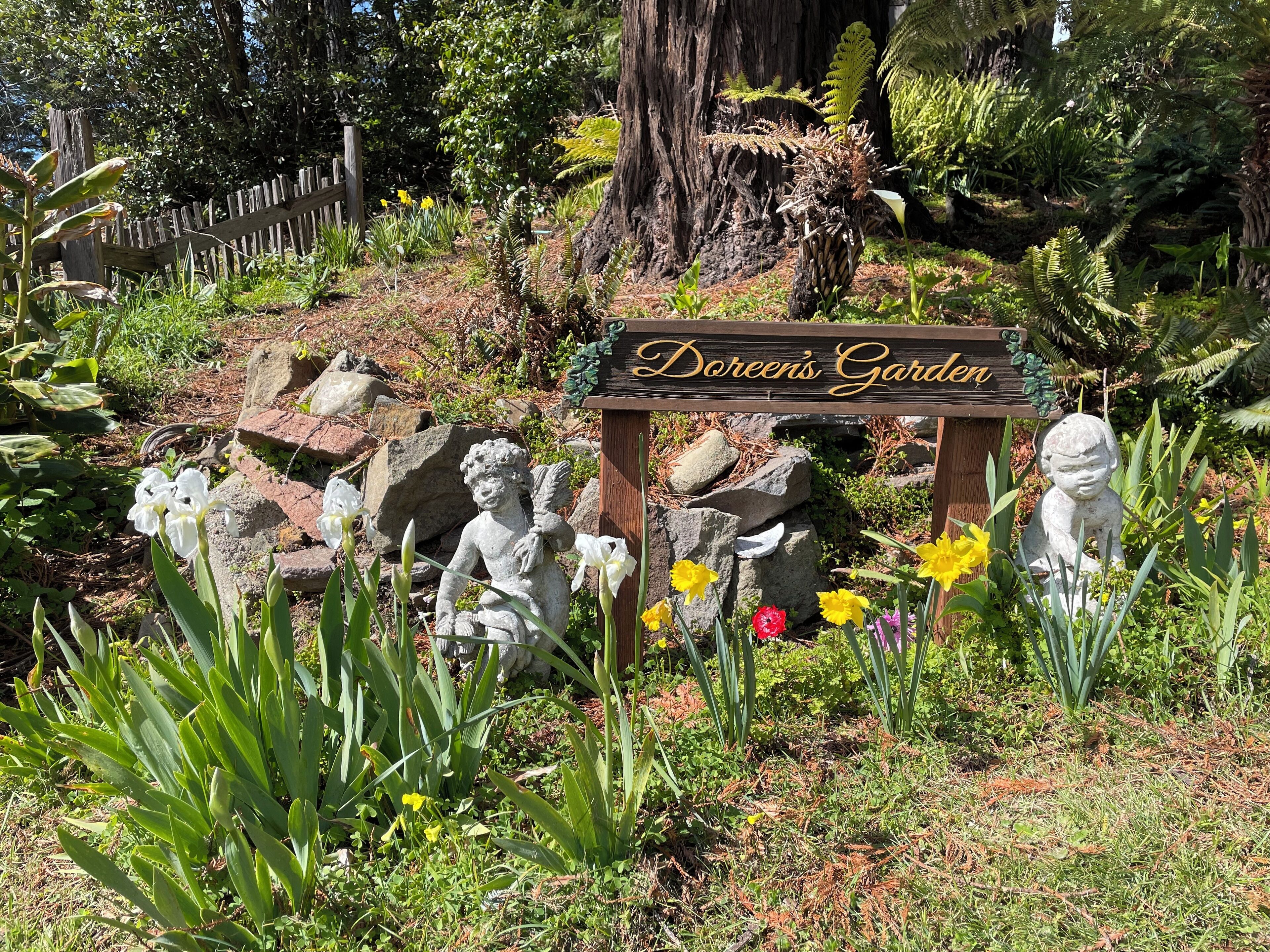 Un jardin agrémenté d’une pancarte en bois sur laquelle on peut lire « Le jardin de Doreen », entouré de diverses plantes et de statues de jardin.