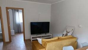 Un saló modern amb televisió de pantalla plana sobre un suport blanc, un sofà groc i una taula de menjador de fusta.