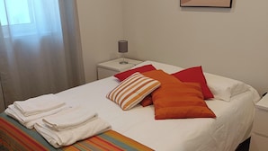 Un llit ben fet amb llençols ratllats, almoines blanques i una almoina colorida. Hi ha una petita taula de nit amb una làmpada, una finestra amb cortines i un quadre penjat a la paret.