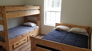 Ein Zimmer mit zwei Holzbettgestellen, eines davon als Etagenbett und das andere mit einem Einzelbett. Ein Fenster sorgt für Tageslicht.