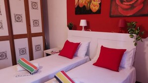 A hotel room with two beds, each with white bedding and red pillows. There are decorative pillows and a striped towel on one bed. The room has a red accent wall with a floral painting and a potted plant. - Bnbook-Appartamento Bilocale Il Geco (Olgiate Olona)