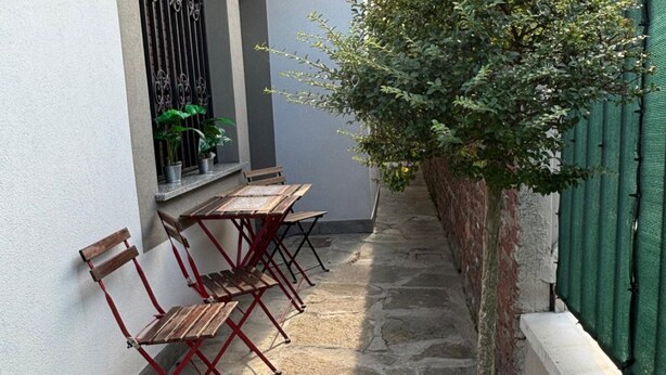 A narrow outdoor corridor with stone flooring, a green fence, and a row of wooden benches with red metal frames. - Bnbook-Appartamento Bilocale Il Geco (Olgiate Olona)