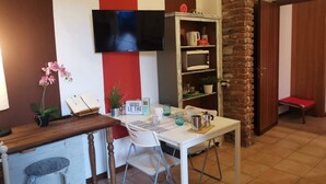 A dining area with a table, chairs, a television, a bookshelf, and a brick pillar. - Bnbook-Appartamento Bilocale Il Geco (Olgiate Olona)