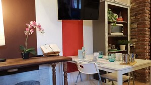 A kitchen with a wooden countertop, a white chair, a white table, a TV mounted on the wall, and a brick wall. - Bnbook-Appartamento Bilocale Il Geco (Olgiate Olona)