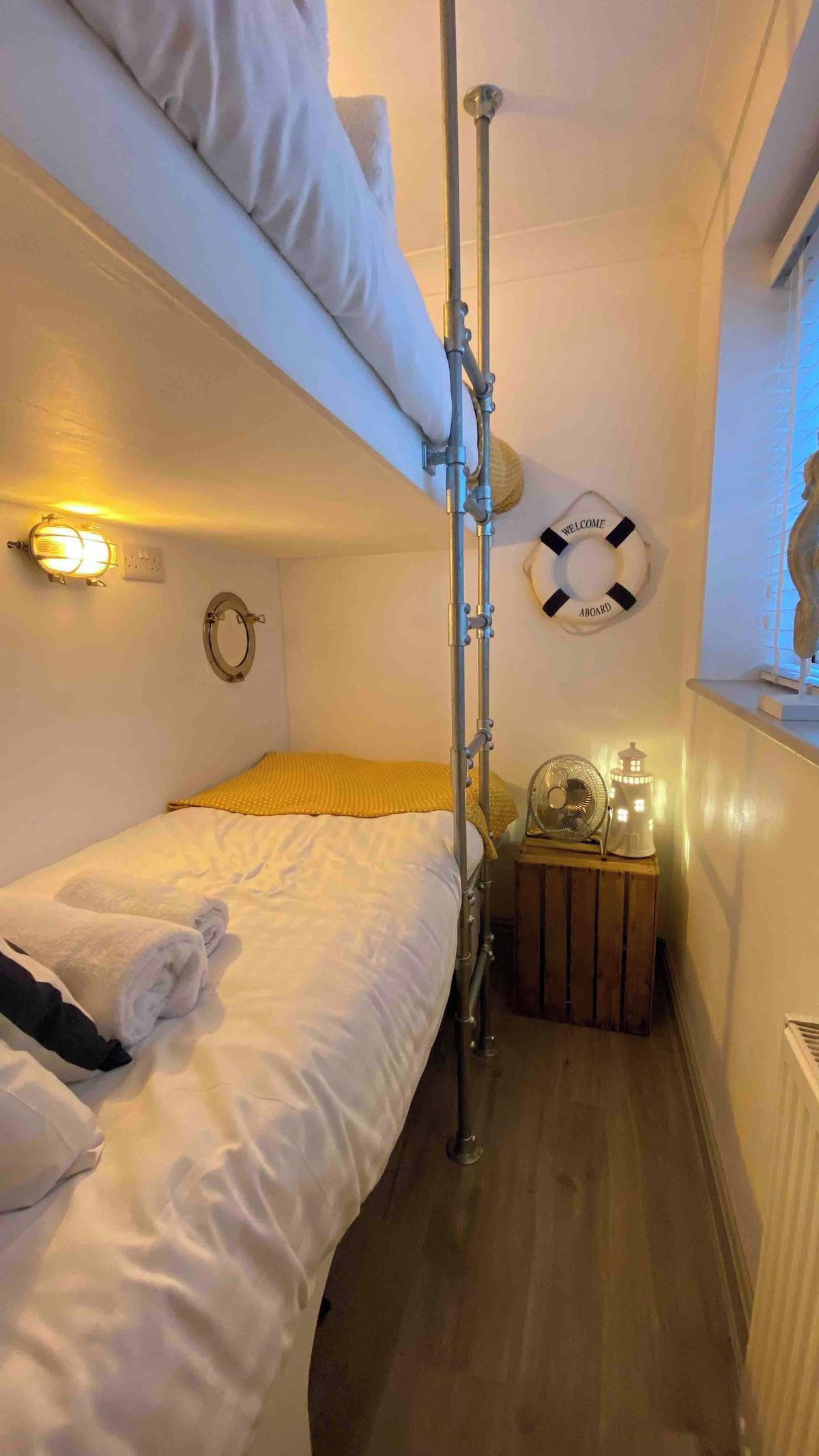 A narrow cabin with bunk beds, a small table with a fan and a lantern, and a life buoy on the wall.