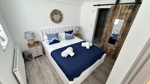 A bedroom with a white bed, blue bedding, and a wooden headboard. There is a bedside table with a lamp and a mirror. - St James Rest - sleeps 4 guests  in 2 bedrooms (St Ives)