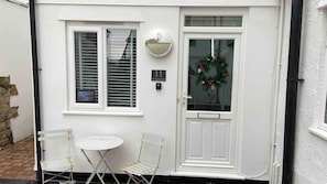 A white exterior with a door, two windows with blinds, a small table with chairs, and a wreath on the door. - Stylish Refurb Apartment with King & Bunk Cabin (St Ives)