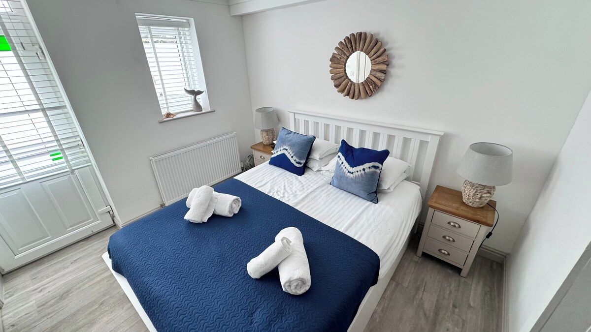 Een slaapkamer met een groot bed, een wit hoofdeinde, twee nachtkastjes met lampen, een spiegel en een raam met jaloezieën.