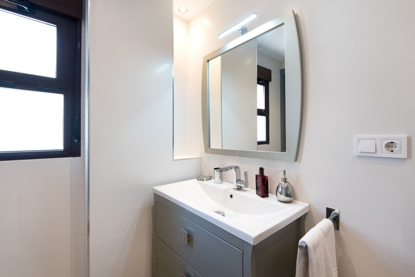 Un baño moderno con tocador blanco, un espejo grande y una ventana.
