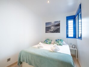 Une chambre à coucher avec un lit, une fenêtre avec des volets bleus, un tableau accroché au mur et une table de chevet sur laquelle se trouve un cadre photo.