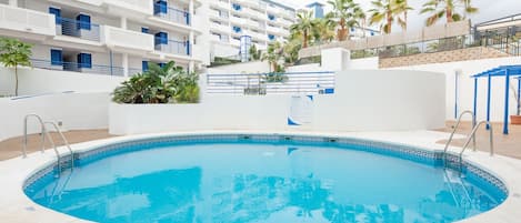 Uma piscina com esquema de cores azul e branco, rodeada por um edifício branco com varandas e palmeiras.
