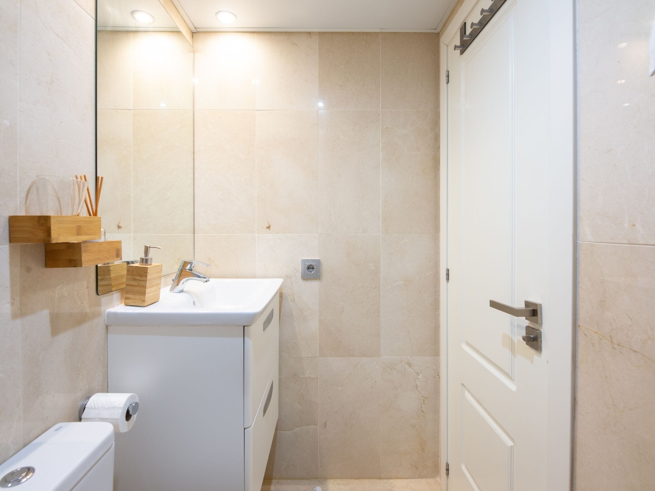 A bathroom with a toilet, sink, and mirror. There is a wooden shelf with toiletries and a towel dispenser.