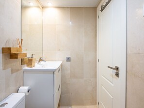 Un bagno con WC, lavandino e specchio. Si vede uno scaffale in legno con articoli da toilette e un distributore di asciugamani.