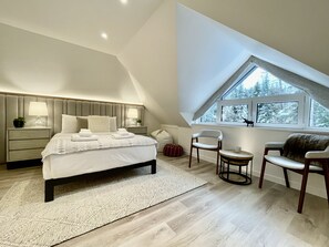 Un dormitorio con una cama grande, mesitas de noche, una silla, una pequeña mesa y una ventana con vistas a árboles.