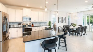 A modern kitchen with white cabinets, stainless steel appliances, a black granite island, and a dining area with a chandelier. - Golden Bear Oasis | 6 Bed, 5.5 Bath Villa, Private Pool, Game Room, Themed Kids' Rooms (Kissimmee)