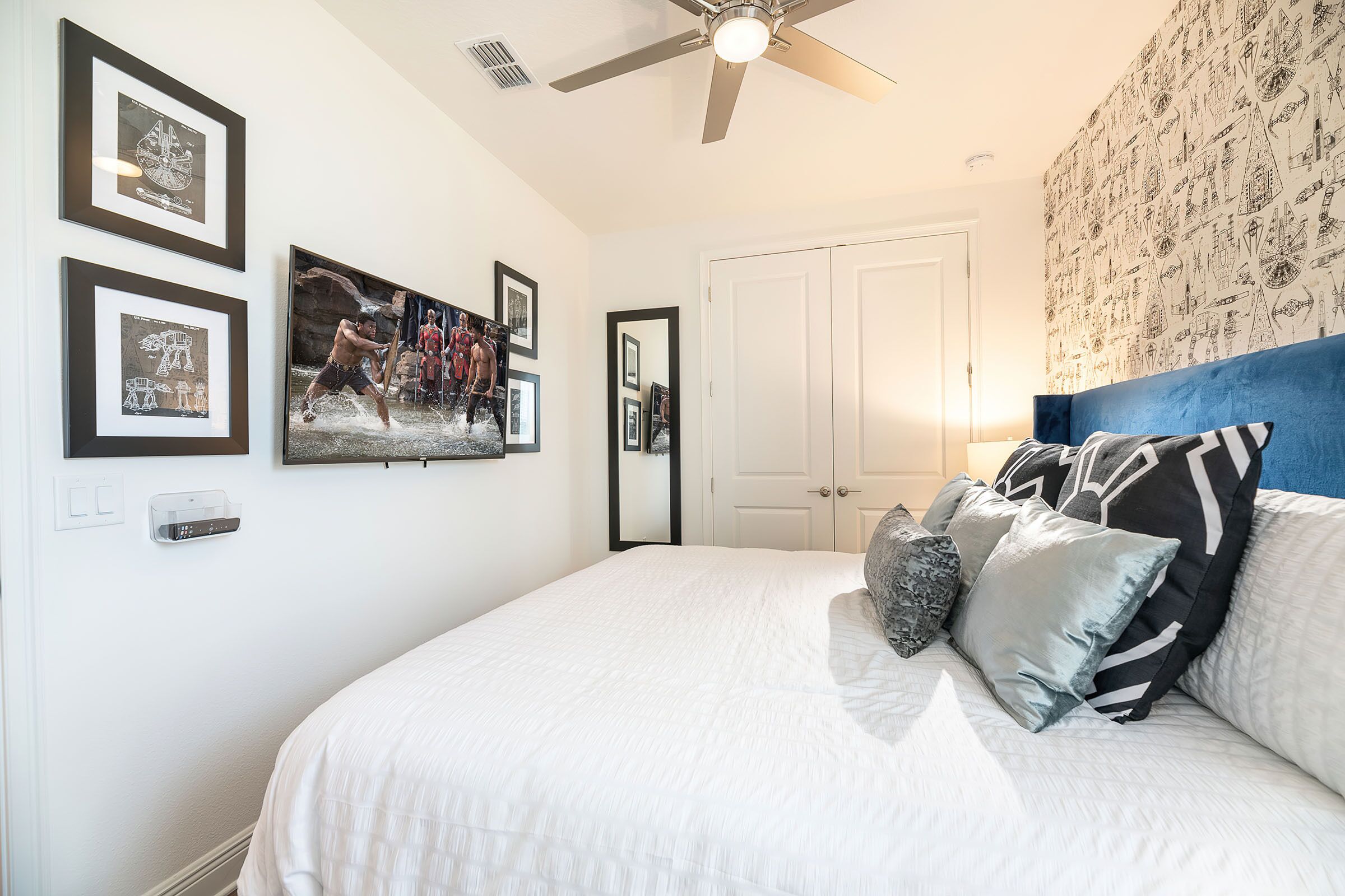 Une chambre à coucher avec un lit, des oreillers, un ventilateur de plafond, des œuvres d’art encadrées et une porte.