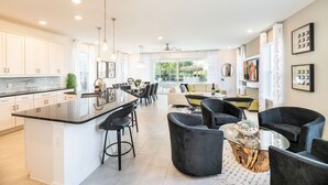 A modern kitchen with a central island, bar stools, and a dining area with a sofa and chairs. - Golden Bear Oasis | 6 Bed, 5.5 Bath Villa, Private Pool, Game Room, Themed Kids' Rooms (Kissimmee)