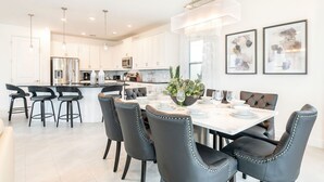 A modern kitchen with white cabinets, a central island, and a dining area with black chairs and a white table. - Golden Bear Oasis | 6 Bed, 5.5 Bath Villa, Private Pool, Game Room, Themed Kids' Rooms (Kissimmee)
