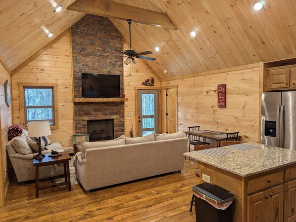 A cozy cabin interior with a stone fireplace, wooden walls, a ceiling fan, a TV, and a kitchen area.