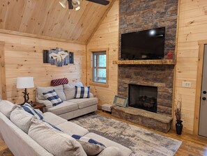 A cozy cabin interior with a stone fireplace, a large flat-screen TV mounted above it, and a comfortable seating area with a sofa and a small side table with a lamp. - Cute Cabin 10mins from Blue Ridge, wifi, hot tub Hiking and waterfalls near (Blue Ridge)